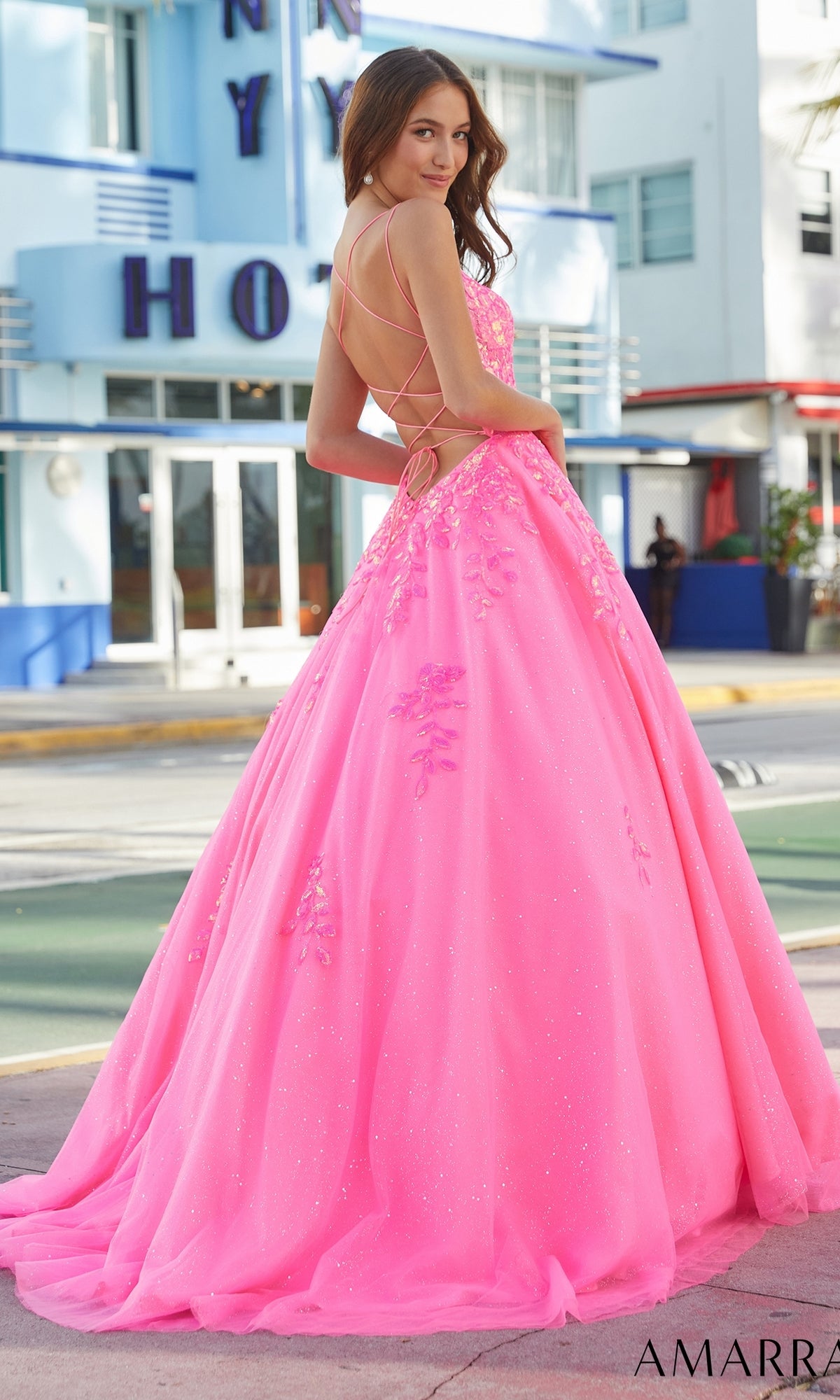 A woman poses outdoors in the Amarra Long Prom Dress 88574, a bright pink gown with floral appliqués, glitter tulle full skirt, strappy open back, and a corset bodice for an elegant touch.
